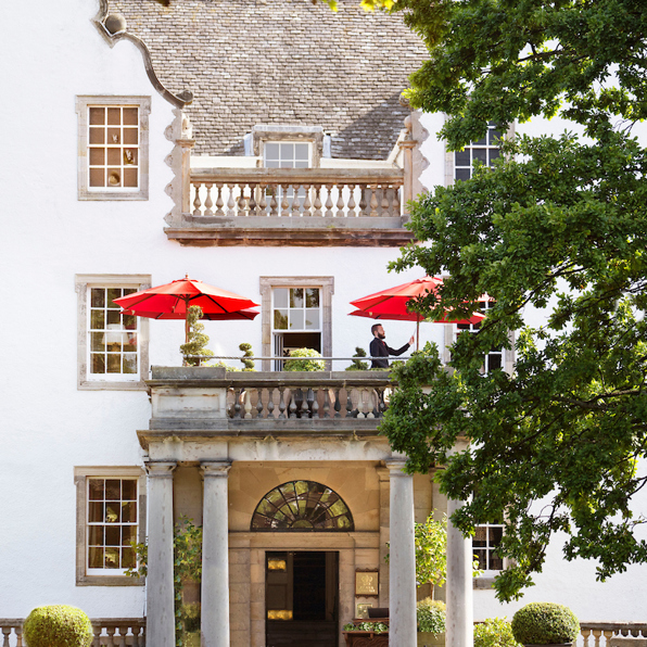 A staff member adjusts a vibrant red parasol on the stone balcony of Prestonfield House, a historic luxury 5-star hotel in Edinburgh, featuring white architectural facades and lush greenery.